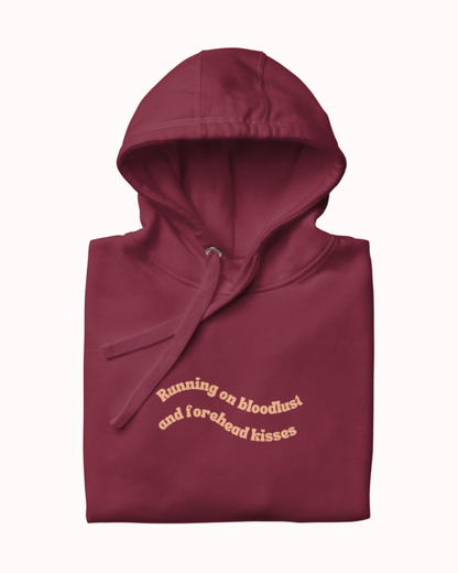 Folded Maroon hoodie featuring the ironic phrase "Running on bloodlust and forehead kisses". Perfect for fans of funny merchandise, bold humor, and witty sarcasm.