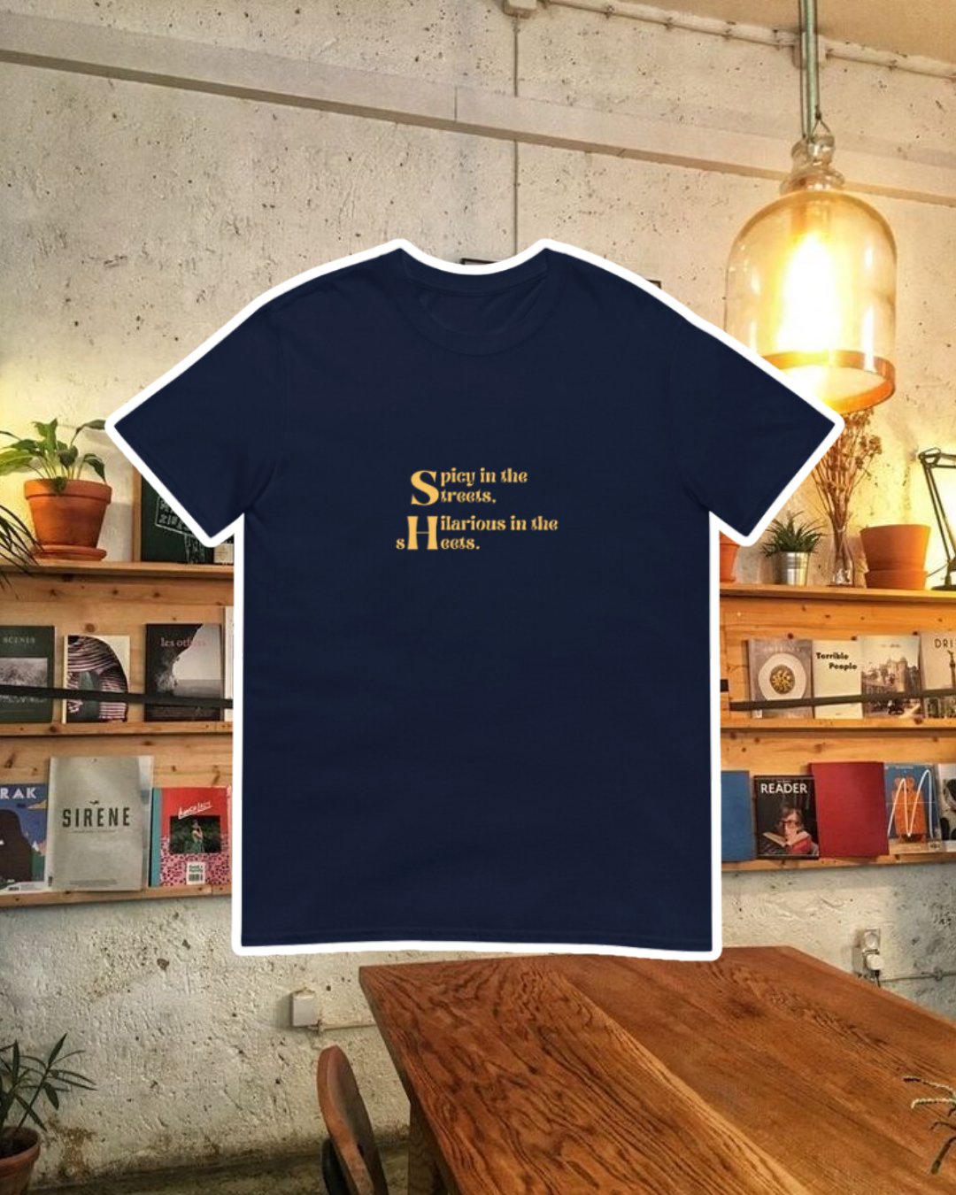 Navy T-Shirt with the phrase 'Spicy in the streets, Hilarious in the sheets' on the chest, displayed against a vibrant café-style backdrop with wood chairs and colorful cozy decoration. A playful and bold design for fans of funny merchandise and sarcasm.