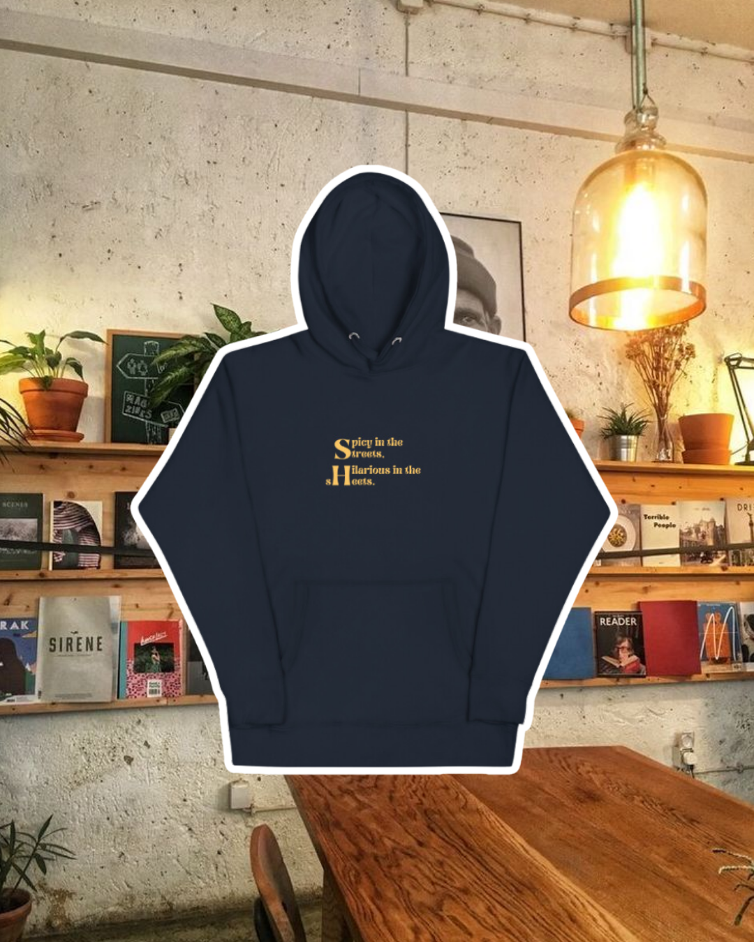 Navy blazer hoodie with the phrase 'Spicy in the streets, Hilarious in the sheets' on the chest, displayed against a vibrant café-style backdrop with wood chairs and colorful cozy decoration. A playful and bold design for fans of funny merchandise and sarcasm.