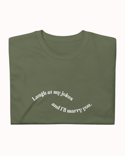 forest green T-Shirt with the phrase 'Laugh at my jokes and I will marry you' on the chest, displayed against a vibrant café-style backdrop with a pink counter and cozy decoration with plants. A playful and bold design for fans of funny merchandise and sarcasm.