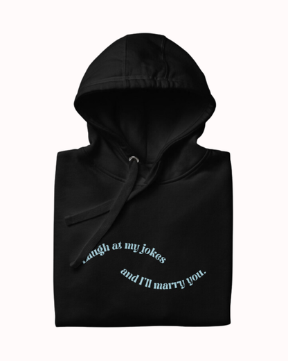 Folded black hoodie featuring the ironic phrase 'Laugh at my jokes and I'll marry you'. Perfect for fans of funny merchandise, bold humor, and witty sarcasm.