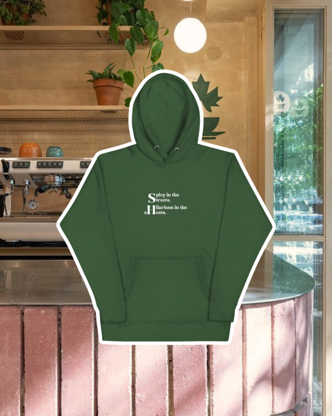 Forest green hoodie with the phrase 'Spicy in the streets, Hilarious in the sheets' on the chest, displayed against a vibrant café-style backdrop with a pink counter and cozy decoration with plants. A playful and bold design for fans of funny merchandise and sarcasm.