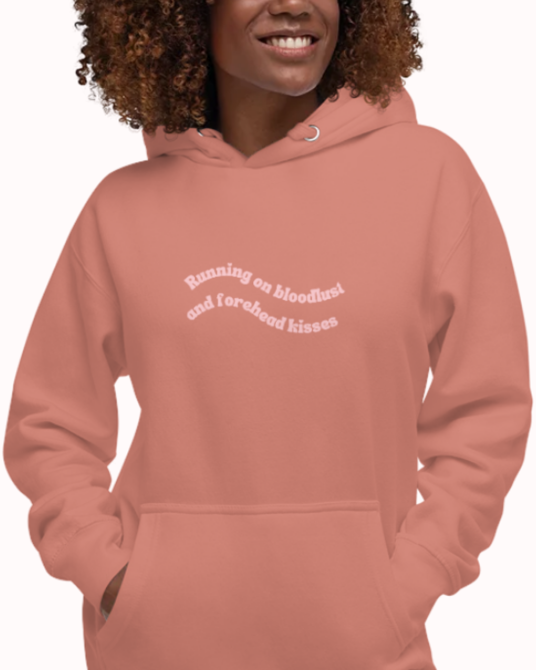Front view of a Dusty Rose hoodie worn by a smiling woman, featuring the phrase "Running on bloodlust and forehead kisses" on the chest and back. Perfect for fans of funny merchandise, bold humor, and witty sarcasm.