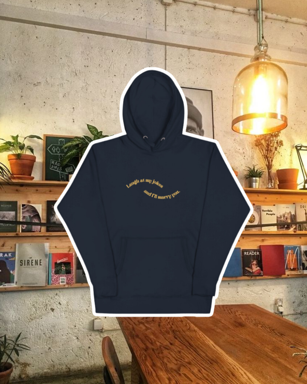 Navy blazer hoodie with the phrase 'Laugh at my jokes and I'll marry you' on the chest, displayed against a vibrant café-style backdrop with wood chairs and colorful cozy decoration. A playful and bold design for fans of funny merchandise and sarcasm.