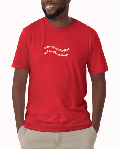 Front view of a Red t-shirt worn by a smiling man, featuring the phrase "Running on bloodlust and forehead kisses" on the chest and back. Perfect for fans of funny merchandise, bold humor, and witty sarcasm.